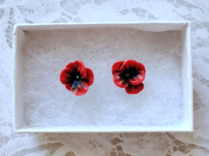 Red and black flower shaped earrings with green metallic centers inside a white paper jewelry box