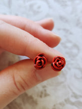 Load image into Gallery viewer, A pair of red and black rose flower earrings held between finger and thumb.
