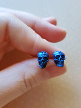 Load image into Gallery viewer, A close of picture of metallic  sapphire colored skull earrings held between finger and thumb. 
