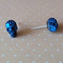 Load image into Gallery viewer, A close of picture of metallic  sapphire colored skull earrings displayed on a pale orange background with white polka dots.
