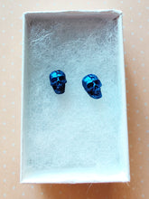 Load image into Gallery viewer, A close of picture of metallic  sapphire colored skull earring in a white paper jewelry box.
