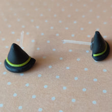 Load image into Gallery viewer, A pair of black witches hats with yellow bands made into flat backed stud earrings. Earrings are displayed on a pale orange background with white polka dots. 
