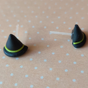 A pair of black witches hats with yellow bands made into flat backed stud earrings. Earrings are displayed on a pale orange background with white polka dots. 