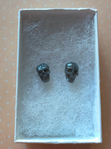 Grey colored skull stud earrings that shimmer silvery in the light inside a white paper jewelry box.  