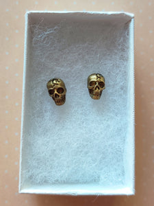 Faux aged gold skull earrings attached to plastic posts inside a white paper jewelry box. 