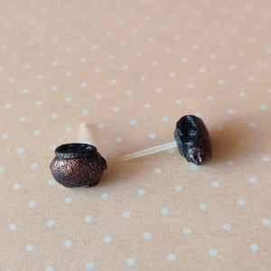 Two miniature flat backed cauldron earrings in black, brushed with a bronze colored metallic pigment. Earrings are displayed on a pale orange background with white polka dots.