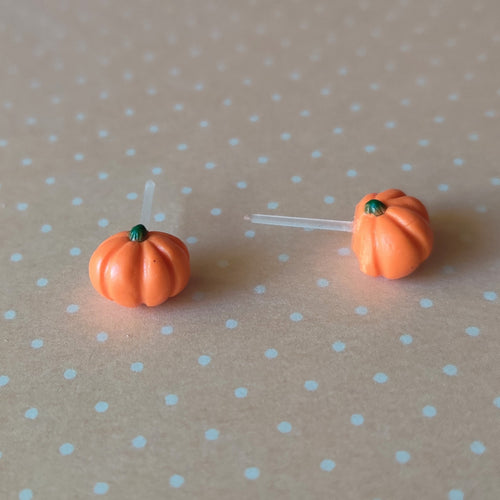A pair of flat backed, side view, orange pumpkin earrings sitting on pastel orange and white polka dot paper.