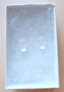 A  pair of clear skull earrings inside a white paper jewelry box. 