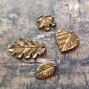 A group of polymer clay leaves showing a gold finish similar to plaited or brushed gold. The leaves are shaped like one leaf each of a red maple, oak, beach, and aspen leaf.