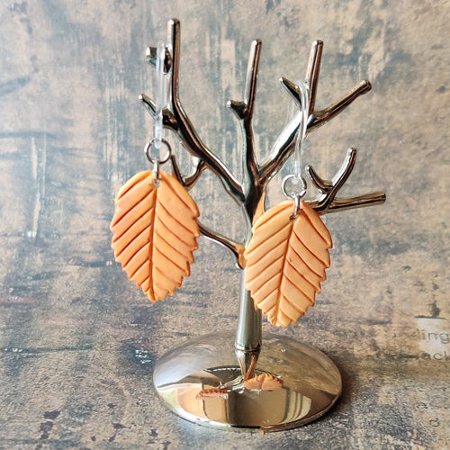 A pair of orange beech leaf shaped polymer clay earrings are displayed hanging from a small, polished silver tree shaped display.