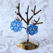 Load image into Gallery viewer, A pair of blue, white, and silver kaleidoscope hexagon earrings hangs from a small polished silver tree stand. 
