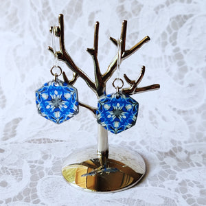 A pair of blue, white, and silver kaleidoscope hexagon earrings hangs from a small polished silver tree stand. 