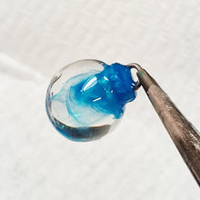 Load image into Gallery viewer, a clear miniature Christmas bauble held by the top with small thin pliers. Swirls of a shimmering blue are inside the clear.
