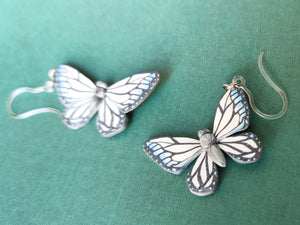 Two white, silver, and blue butterfly earrings with stainless steal jump rings and plastic ear hooks. One is in the foreground is clear and in focus, while the other sits just outside of focal range. 