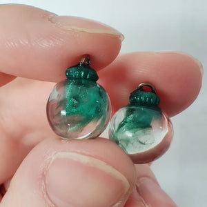 Two miniature Christmas ornament baubles in a clear color with silvery green swirls inside. The are held between finger and thumb. 