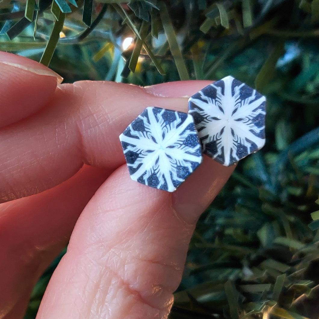 Two hexagon shaped earrings with a snowflake pattern in silver and white. The earrings are held between finger and thumb next to an artificial Christmas tree branch.