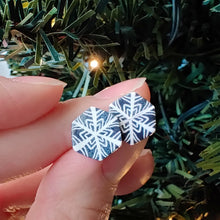 Load image into Gallery viewer, Two hexagon shaped earrings with a snowflake pattern in silver and white. The earrings are held between finger and thumb next to an artificial Christmas tree branch.
