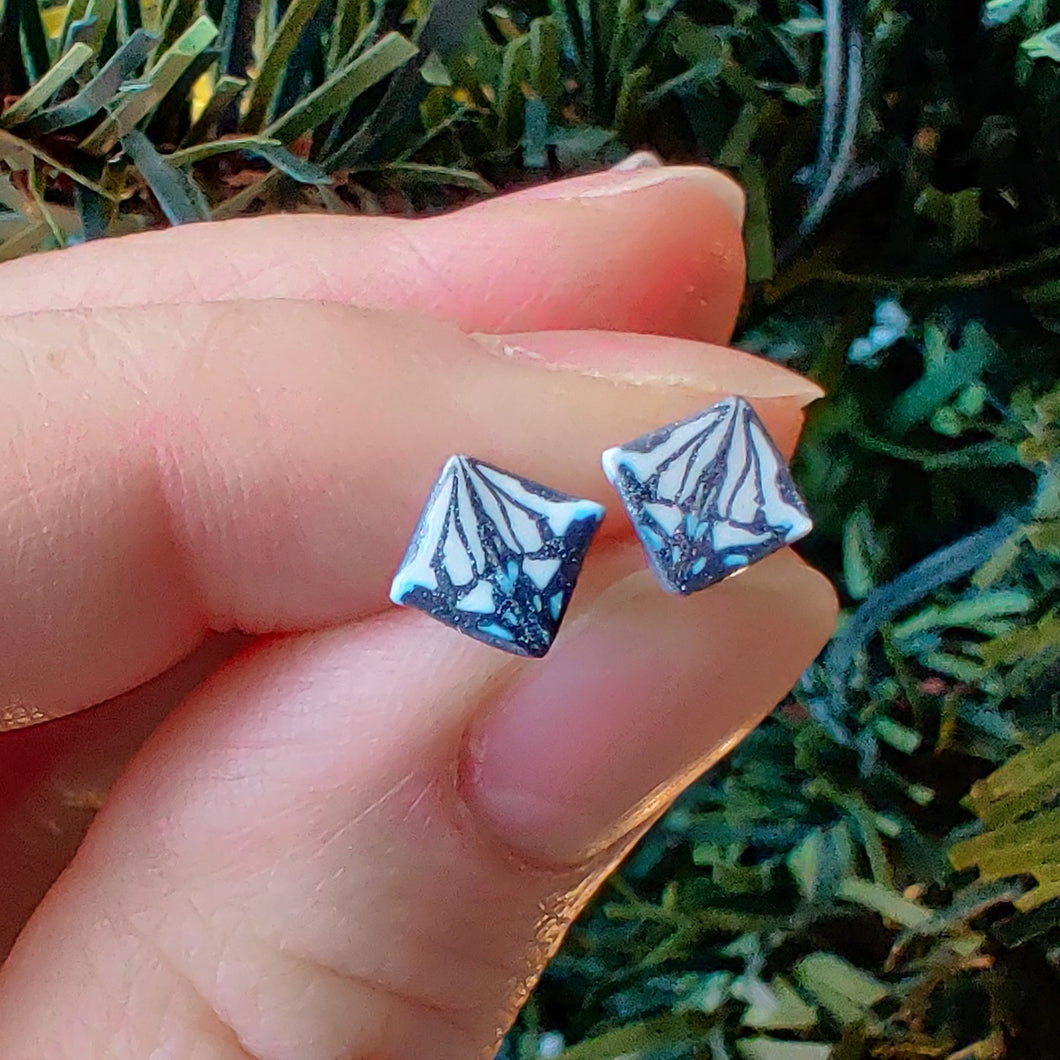 A symmetrical square abstract design of polymer clay earring studs in white, blue and metallic silver held between finger and thumb in front of an artificial Christmas tree.