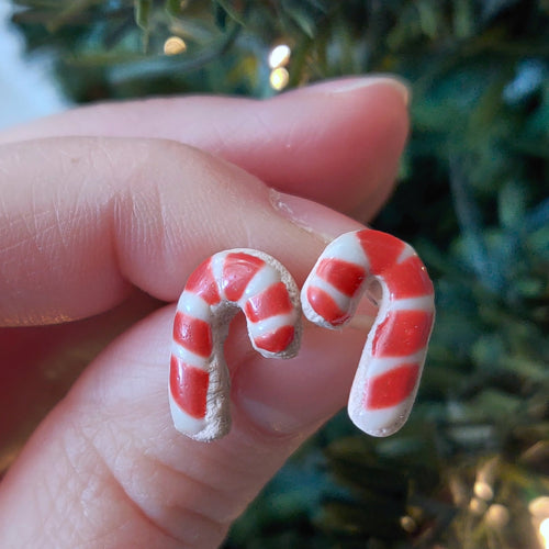 A pair of earrings made to look like sugar cookies decorated as candy canes. The earrings are held between finger and thumb in front of an artificial Christmas tree. 