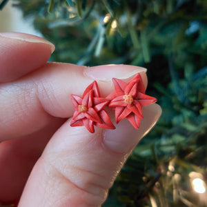 A pair of red Poinsettia earrings held between finger and thumb in front of an artificial Christmas tree.