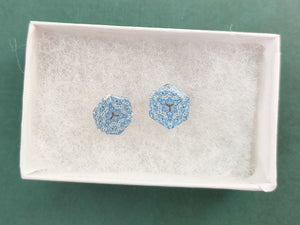 Two hexagon shaped earrings with intricate white, silver and blue patterns in a white paper jewelry box. 