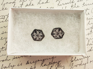 Two hexagon shaped earrings  with a kaleidoscope pattern reminiscent of a snowflake or stained glass. The earrings are inside a white paper jewelry box. 