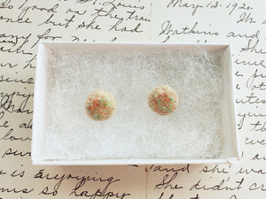 A pair of earrings made to look like sugar cookies decorated with green and red sugar sprinkles. The earrings are inside a white paper earring box. 