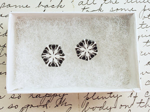 Two hexagon shaped earrings with a snowflake pattern in silver and white. They are in a white paper jewelry box. 