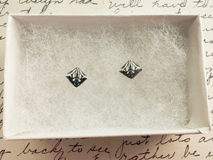 A symmetrical square abstract design of polymer clay earring studs in white, blue and metallic silver held between finger and thumb inside a white paper jewelry box.