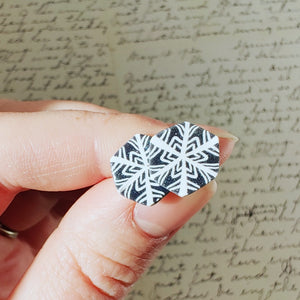 Two hexagon shaped earrings with a snowflake pattern in silver and white. The earrings are held in bright sunlight and have small silver sparkles in the grey areas. 