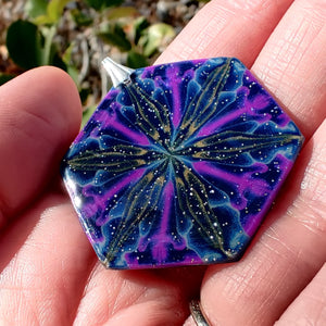 A large hexagonal pendant held in hand in the sunlight. The pendent is dark blue with purple, gold, and light blue colors radiating out from the center. The light reflects off of tiny gold and blue sparkles and small silver glitter. The whole pendant has a shiny gloss finish. 