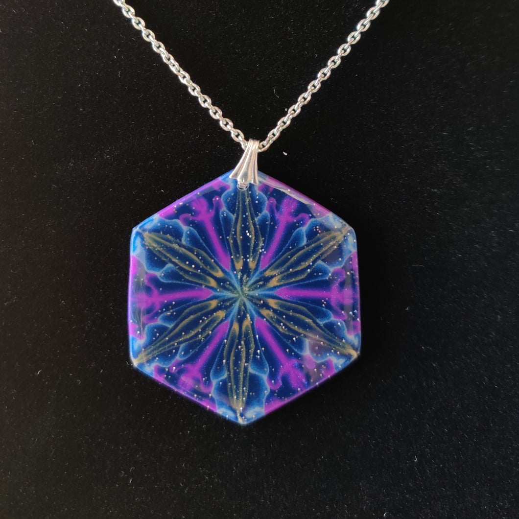 A large hexagonal pendant hanging from a silver colored chain and bail. The pendent is dark blue with purple, gold, and light blue colors radiating out from the center. It is displayed with a black velvet background. 