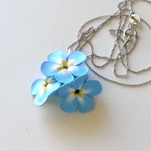 A small pendant attached to a silver colored chain and bail. The pendant is a cluster of three imitated Forget Me Not flowers. The flowers are pale blue and fade to white toward the center. Each petal ends in yellow at the middle and the center is black. The pendant is displayed on a white ceramic background. 