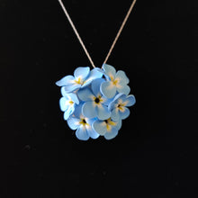 Load image into Gallery viewer, A large pendant hanging from a silver colored chain displayed on a black velvet background. The pendant is a round cluster of seven Forget Me Not imitation flowers made of polymer clay. Each of the flower petals is blue at the edges, fades to white, and ends in yellow in the middle. The center of the flowers is black. 
