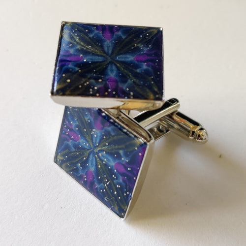 A pair of silver colored cufflinks with a square bezel on top. The bezel is filled with polymer clay with a shiny gloss finish. The clay is dark blue with veins of gold, purple, and glow in the dark clay radiating from the center. 
