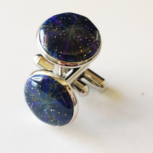 Load image into Gallery viewer, A pair of silver colored cufflinks with a bezel on top. The bezel is filled with polymer clay with a shiny gloss finish. The clay is dark blue with veins of gold, purple, and glow in the dark clay radiating from the center. 
