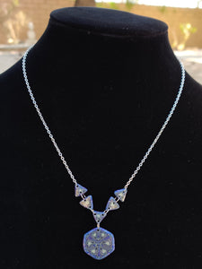 A silver colored chain neckalce connects to a series of 5 triangles in the center of a neckalce. From the center triangle hangs a hexagonal pendant. The necklace is blue, purple, and glow in the dark. 