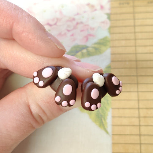 Two small bunny butt shaped earrings between finger and thumb. They are brown with pink foot pads, three toes, and a white tail.