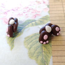Load image into Gallery viewer, Two small bunny butt shaped earrings resting on a  flowered background. They are grey with pink food pads, three toes, and a white tail.
