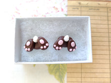 Load image into Gallery viewer, Two small bunny butt shaped earrings sitting inside a white paper jewelry box. They are brown with pink foot pads, three toes, and a white tail.
